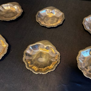 May include: Six small, gold-colored, decorative dishes shaped like seashells. The dishes are arranged in a semi-circle on a black surface.