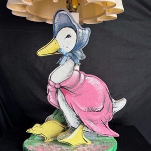 May include: A wooden lamp base with a painted design of a duck wearing a pink dress and blue bonnet, holding a yellow duckling. The lamp has a white pleated lampshade.