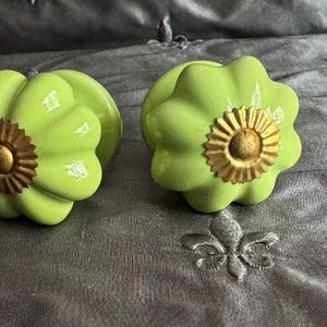 May include: Two lime green cabinet knobs with a flower-like design and a gold-colored center. The knobs are on a gray fabric surface. These decorative knobs can be used for furniture or other home decor projects.