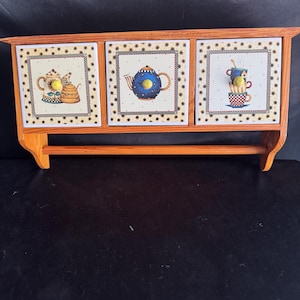 May include: Wooden shelf with three decorative ceramic tiles featuring teapots and teacups. The tiles have a sunflower border and are mounted on a light brown wooden frame with a towel bar.