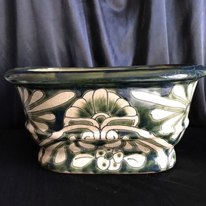May include: A ceramic planter with a wide, oval shape. The planter is decorated with a floral pattern in shades of green and white against a dark blue background. The rim and base are dark blue.