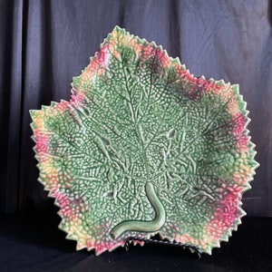 May include: A large green ceramic leaf-shaped serving dish with a raised design. The dish has a pink and yellow border and a green vine detail.