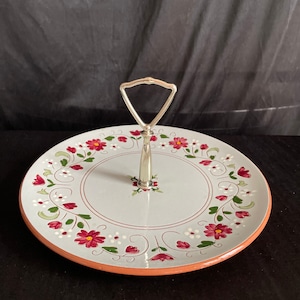May include: A two-tiered serving tray with a silver handle. The tray features a light blue surface with a floral design of red flowers, green leaves, and white accents. The rim is a terracotta color.
