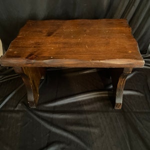 May include: A small, rectangular wooden stool with a dark brown stained finish. The top surface features a natural, uneven live edge along the sides, and the stool is supported by two sturdy, curved wooden legs on either side of the base.