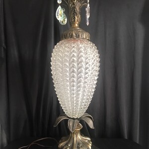 May include: Vintage table lamp with a clear, textured glass shade shaped like a pinecone. The lamp features a brass-colored base and accents, with decorative crystal pendants. The base is black with ornate detailing.