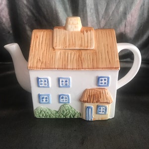 May include: A white ceramic teapot shaped like a house, featuring a tan roof and chimney, blue window frames, and a small door. Green bushes are at the base of the house. The spout and handle are white.