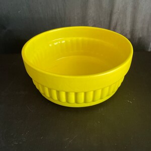 May include: A bright yellow ceramic bowl with a ribbed design around the base. The bowl has a smooth, glossy finish and a simple, rounded shape. This bowl is suitable for serving or as a decorative piece.