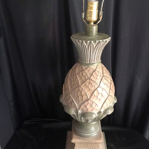 May include: A decorative table lamp with a pineapple-shaped base. The lamp features a beige and green color scheme, with a textured surface on the pineapple design. The lamp has a brass-colored bulb fixture and a light bulb.