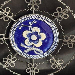 May include: A decorative plate with a blue and white floral design. The center of the plate features a blue background with a white flower. The plate is surrounded by a silver metal wire frame with flower-shaped accents.