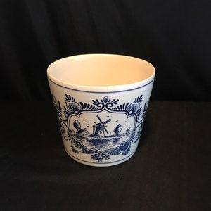 Puede incluir: Maceta de cerámica blanca con un diseño de molino de viento holandés azul y blanco. La maceta presenta una escena de paisaje detallada con molinos de viento, árboles y agua. El borde y la base tienen patrones decorativos azules.