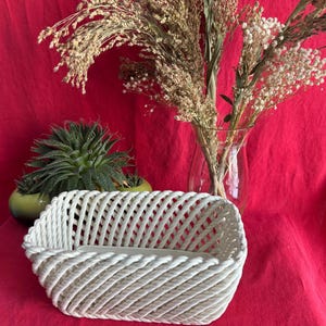 May include: A white woven basket with a rectangular shape. The basket is made of thick, twisted rope-like material. The background features a red fabric, a glass vase with dried flowers, and several small green potted plants.