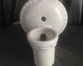 Op de afbeelding: Een witte, aan de muur te bevestigen tandenborstelhouder van keramiek met een geschulpte rand en een klein gaatje. De houder heeft een cilindrische beker en een cirkelvormige achterplaat. Eenvoudig, klassiek ontwerp voor de badkamer.