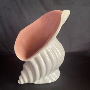 May include: A white ceramic seashell vase with a pink interior. The vase is shaped like a conch shell and has a smooth, glossy finish.