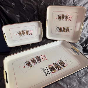May include: A set of three white serving trays with gold-toned rims and cream-colored handle grips. Each tray features a graphic print of a hand of playing cards in the center, enclosed by a thin red rectangular border on the white surface.