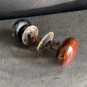 May include: Two antique door knobs. One knob is black and the other is brown. The knobs are oval-shaped with a glossy finish. The hardware is made of metal with a rusted appearance. The door knobs are on a gray surface.