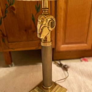 May include: A brass table lamp with a pleated lampshade. The lamp features a ram's head design on the stem and a square base. The lamp's stem is a fluted column. The lampshade is a cream color.