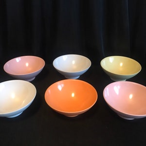 Puede incluir: Seis cuencos de cerámica de colores pastel. Los cuencos son en tonos rosa, blanco, amarillo y naranja. Cada cuenco tiene una forma redondeada con una base blanca. Los cuencos están dispuestos sobre una superficie negra.