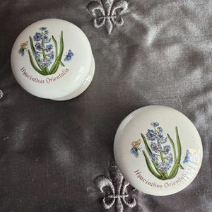 May include: Two white, round decorative ceramic pieces with a floral design. The design features a cluster of blue and purple flowers, green leaves, and butterflies. The text "Hyacinthus Orientalis" is printed on the ceramic.