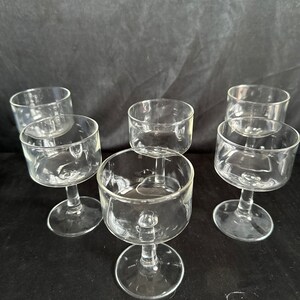 May include: Six clear glass stemmed drinking glasses with a wide bowl and a small base. The glasses are arranged in a circle on a black surface.