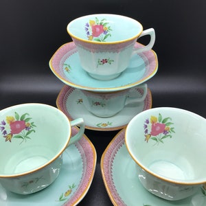 May include: A set of four vintage teacups and saucers. The cups and saucers are light blue with a pink and gold trim. Each cup features a floral design with pink, yellow, and purple flowers. The saucers have a matching pink trim.