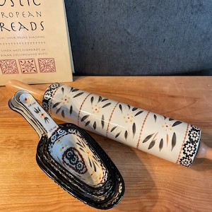 May include: A rolling pin and measuring scoops with a floral design. The rolling pin is white with black, yellow, and brown accents. The measuring scoops are black with a floral pattern. A cookbook is in the background.