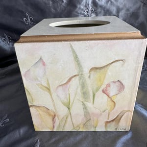 May include: A square tissue box with a floral design. The box features a light beige background with watercolor-style calla lilies in shades of pink, yellow, and green. The top has a rectangular opening and a gold trim. The artist's signature, "BLUM", is visible.