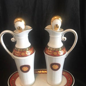 May include: A white ceramic oil and vinegar set with gold accents. The set includes two pitchers with handles and a matching oval tray. Each pitcher has a gold-colored lid and a decorative band with a Greek key design in red and gold.
