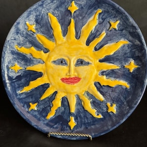 May include: Hand-painted ceramic plate featuring a smiling sun with yellow rays and a red mouth against a dark blue background. The plate is decorated with yellow star accents. A decorative piece for the home.