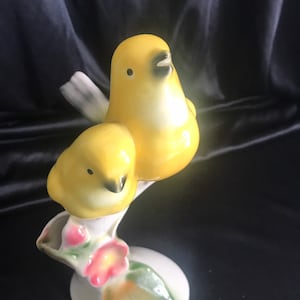 May include: A ceramic figurine of two yellow birds perched on a branch with pink flowers. The birds are painted in a realistic style and have black eyes and beaks.