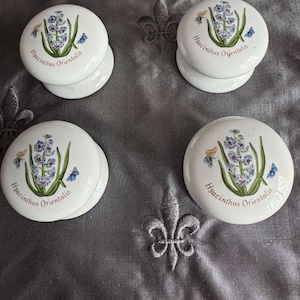 May include: Four white ceramic cabinet knobs with a floral design featuring blue and purple flowers, green leaves, and butterflies. The text "Hyacinthus Orientalis" is printed on each knob.