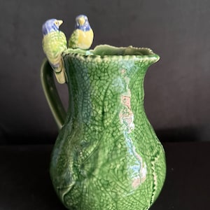 May include: A green ceramic pitcher with a leafy design and two small blue and yellow birds perched on the rim.