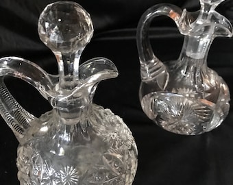 May include: Two clear glass decanters with faceted stoppers and handles. The decanters have intricate floral and starburst patterns etched into the glass. The decanters are on a dark background.