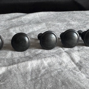 May include: Six black metal knobs with a round, slightly raised, smooth surface. The knobs are arranged in a row on a white surface.