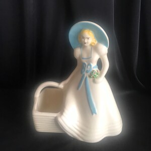 May include: A white ceramic planter shaped like a woman wearing a blue hat and dress. The woman is holding a bouquet of flowers and has a blue ribbon around her waist. The planter has a basket-shaped base.