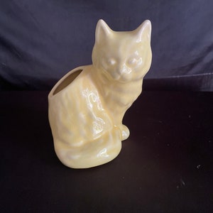 May include: A pale yellow ceramic cat planter. The cat is sitting with its head tilted slightly to the side. The planter has a hole in the back for drainage.
