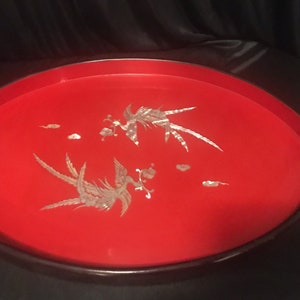 May include: A round, red serving tray with a dark brown rim. The tray features two stylized bird designs and small cloud-like accents in a silver color. The tray is likely made of wood and has a glossy finish.