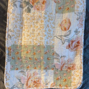Puede incluir: Una pieza de tela acolchada rectangular con un diseño de patchwork de motivos florales en tonos melocotón, verde salvia y blanco. El tejido presenta líneas de costura verticales y un borde fino estampado que enmarca los motivos florales.