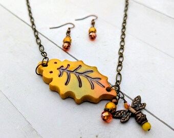 May include: A necklace and earring set featuring a yellow and orange gradient leaf pendant with a dark metal chain. The necklace includes a small metal dragonfly charm and orange crystal beads. The matching earrings have similar beads.