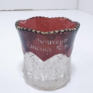 May include: A vintage souvenir glass with a clear, cut-glass base and a burgundy-colored top. The top has the text "Souvenir Lincoln Neb." in silver lettering. The rim is trimmed with a gold and black design.