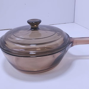 May include: A brown glass saucepan with a matching lid and a long handle. The lid has a small, round knob. The pan is designed for cooking and has a simple, functional design.