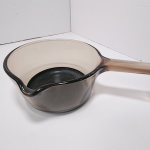 May include: A clear glass saucepan with a long handle. The pan has a dark interior and a light brown tint. The handle is also light brown and extends from the side of the pan. The pan is sitting on a white surface.