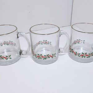 May include: Three clear glass mugs with gold rims and handles. Each mug features a festive design of red ribbons and green holly leaves. The mugs are perfect for holiday beverages.