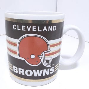 May include: White ceramic mug featuring the Cleveland Browns logo. The mug has a brown background with the team's helmet in the center. The team name is printed in white. Gold bands accent the top and bottom.