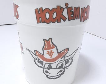 May include: White plastic cup with the words "Hook 'Em Horns" in orange lettering. The cup features a cartoon longhorn wearing a cowboy hat. The cup is a promotional item for the University of Texas.