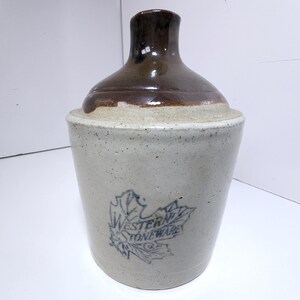 May include: A vintage stoneware jug with a brown top and a light beige body. The jug features a blue leaf design with the words "Western Stoneware" printed on the front. The jug has a cylindrical shape with a rounded top and a narrow neck.