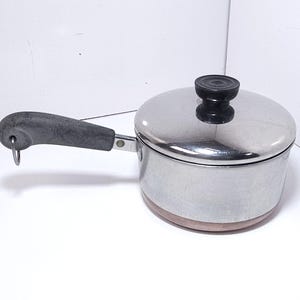 May include: A small, stainless steel saucepan with a lid and a black handle. The pan has a copper-colored bottom and a black knob on the lid. The handle has a hole for hanging. Kitchen cookware.