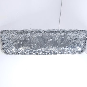 May include: A rectangular, silver-colored metal tray with a detailed relief design of rabbits among foliage. The tray's edges are scalloped, and the rabbits are intricately rendered, creating a decorative and functional piece.