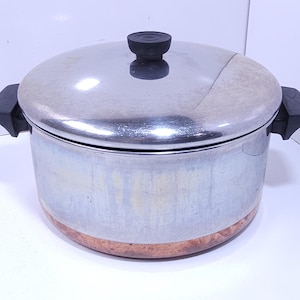 May include: A stainless steel stockpot with a matching lid and black plastic handles. The pot features a polished silver upper body and a textured copper-colored base. The lid has a central black knob and a smooth, reflective metallic surface.
