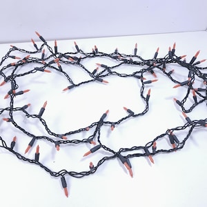 May include: A string of decorative lights featuring small, orange-colored bulbs attached to a black wire. The lights are arranged in a loose, coiled formation, showcasing multiple individual bulb sockets spaced along the dark, flexible electrical cord.