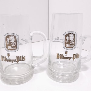 May include: Two clear glass beer mugs with handles. Each mug features a logo with a portrait and the words "Bitburger Pils" in brown. The mugs are empty and appear to be vintage.
